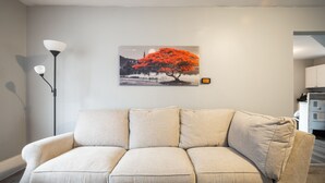 Ein Wohnzimmer mit einem beigen Sofa, einer Stehlampe und einem an der Wand hängenden Bild von einem Baum.