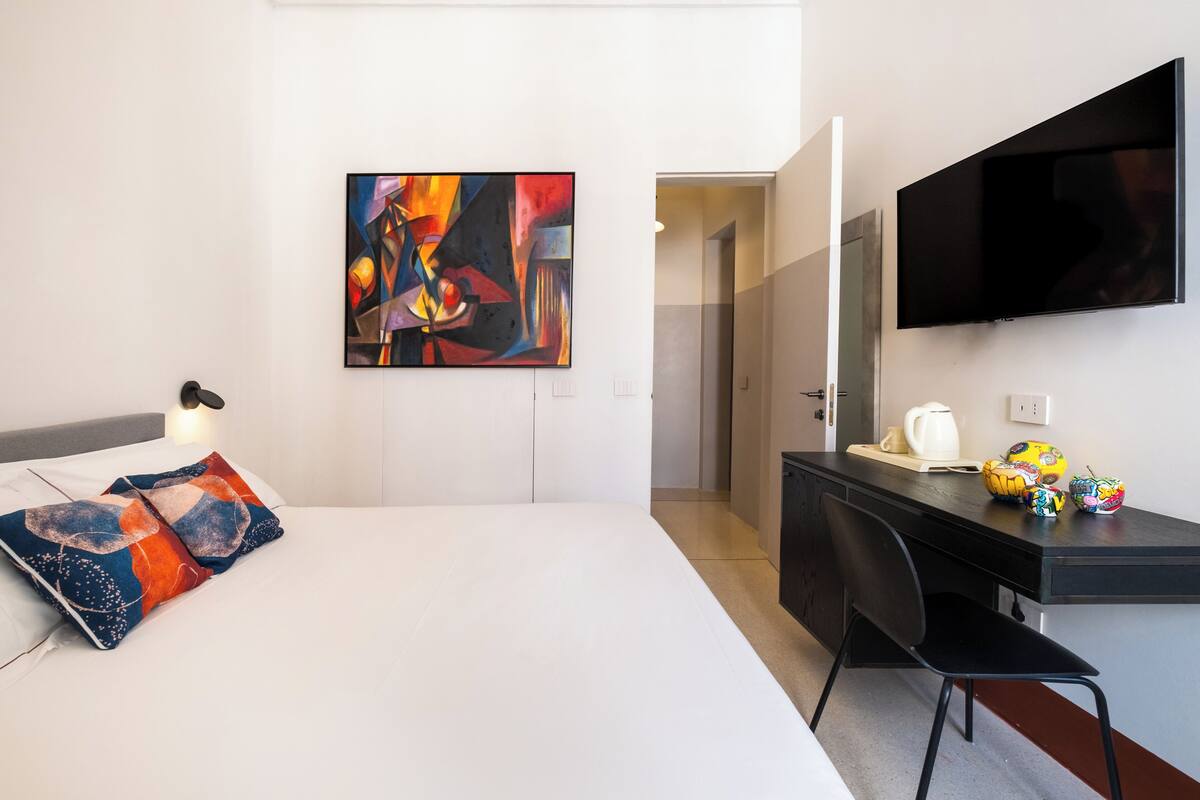 Una habitación de hotel moderna con una cama, un escritorio, una silla, un televisor de pantalla plana y un cuadro abstracto y colorido en la pared.