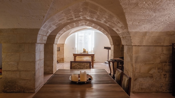 Un corredor de piedra en arco con una mesa y sillas de madera, que conduce a un ventanal con luz natural.