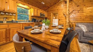 Interior de una cabaña de madera con una mesa de comedor preparada para cuatro comensales, zona de cocina y un rincón de descanso acogedor con sofá.