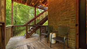 Una terraza de madera con una escalera que conduce a un espacio cubierto, rodeada por un muro de ladrillo, con mobiliario de exterior y vistas a un bosque.