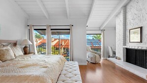 A bedroom with a large bed, a fireplace, and a view of a red-roofed building through a sliding glass door. - Carlsbad Hideaway. Pool, Spa, BBQ. Walking to restaurants/shops (Carlsbad)