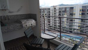 A balcony with a small table and chairs, a grill, and a view of a swimming pool and buildings.