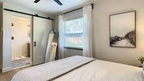 A bedroom with a bed, a ceiling fan, a window with curtains, a door leading to a bathroom, and a painting on the wall. - Cypress Inn - Modern home close to Siesta Key (Sarasota)