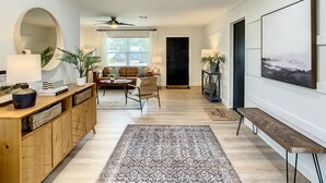 A spacious living area with a sofa, coffee table, and a large mirror. There is a ceiling fan, a window with blinds, and a door leading to another room. - Cypress Inn - Modern home close to Siesta Key (Sarasota)