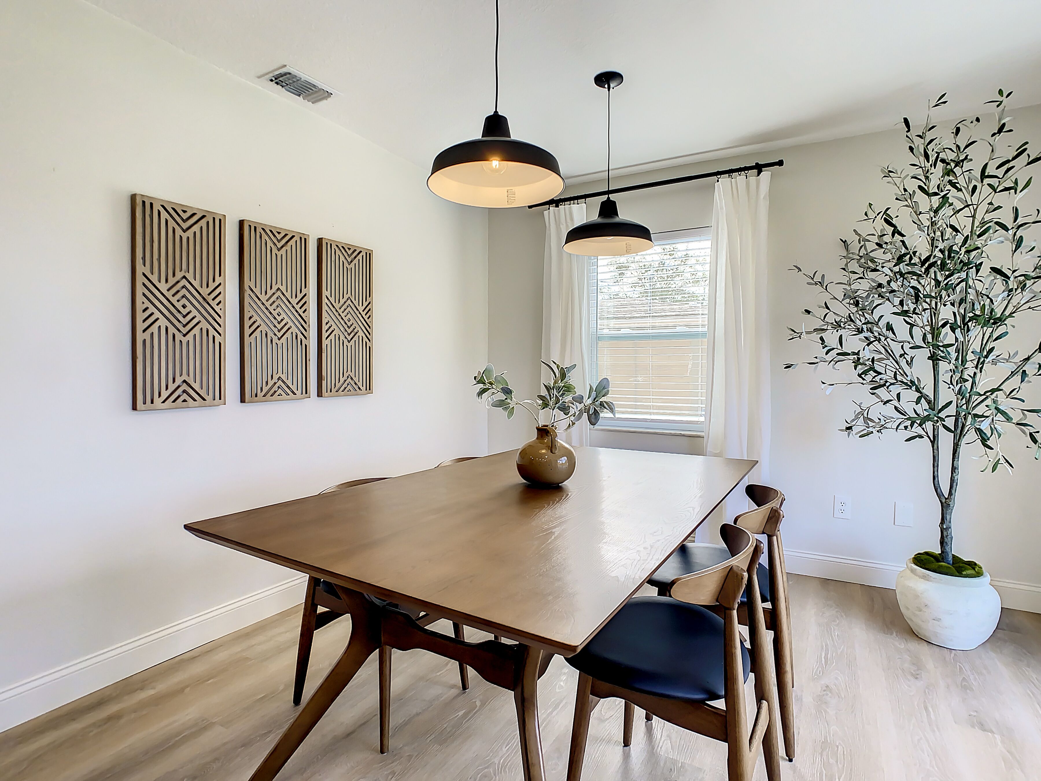 Un comedor con una mesa de madera, sillas, luces colgantes, decoración en la pared y una planta en maceta.