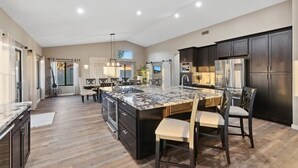 A modern kitchen with dark cabinetry, a large island with a marble countertop, and a dining area with chairs. - Luxury Home, Beautiful Neighborhood, Spacious for Family, with Pool/Spa.  (Glendale)