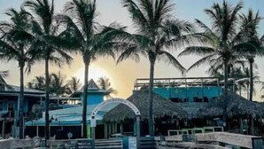 A beachfront walkway with palm trees, a thatched-roof structure, and a building with a sign. - Malvia’s Oasis- Cozy home with a Private Pool (Englewood)