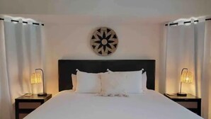 A neatly made bed with white linens, two bedside lamps, and a decorative wall piece above the headboard. - Malvia’s Oasis- Cozy home with a Private Pool (Englewood)