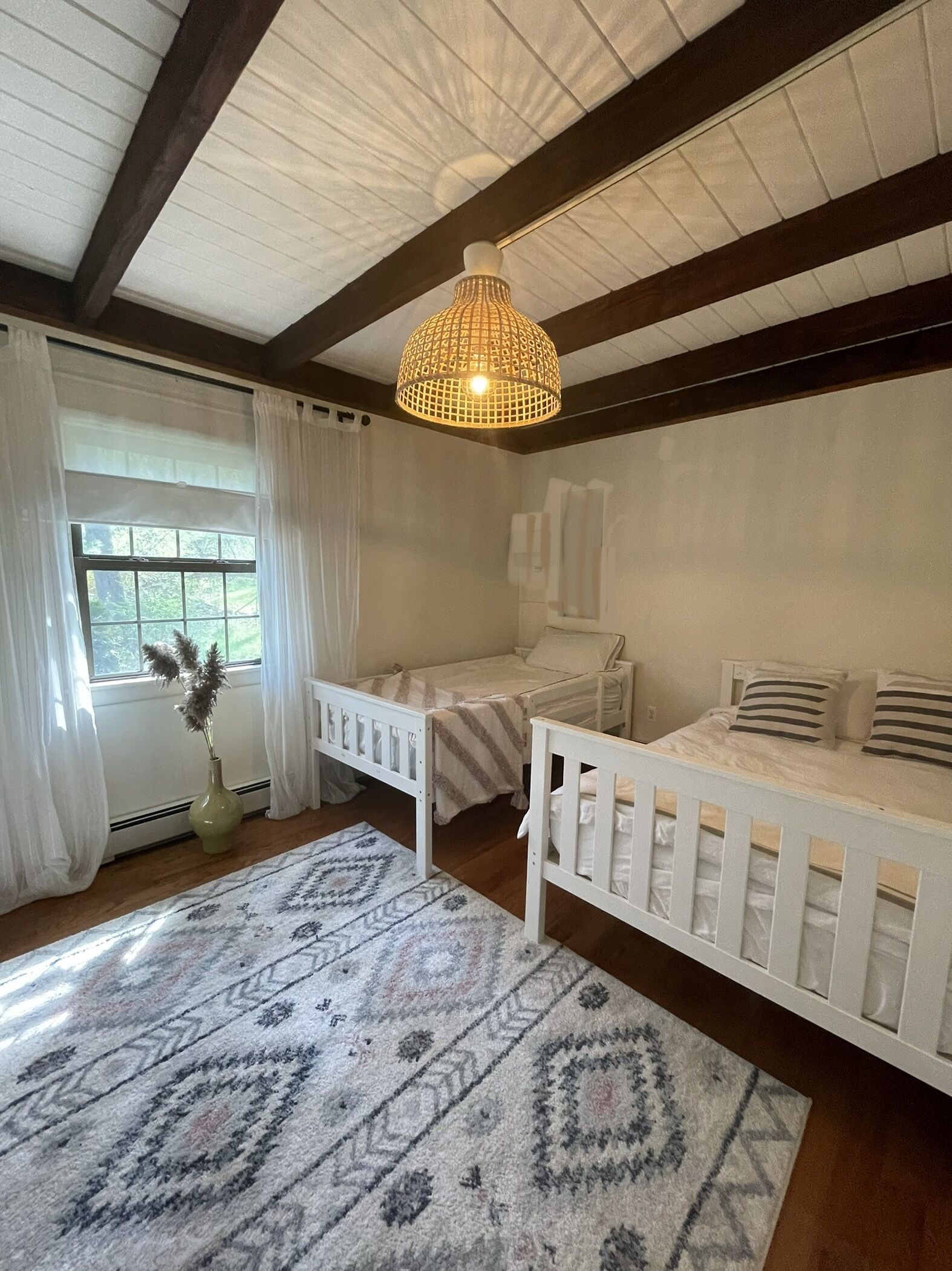 Une pièce avec deux berceaux blancs, un tapis à motifs, un luminaire suspendu et une fenêtre avec des rideaux.