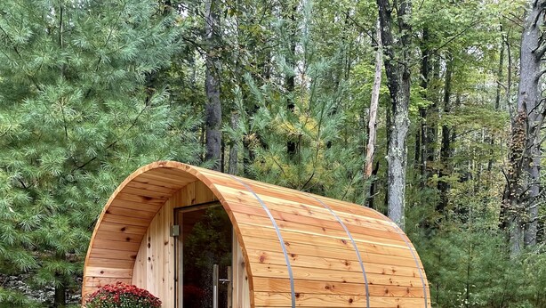 Una cabaña cilíndrica de madera con una puerta de cristal, un pequeño rincón de descanso y una maceta afuera.