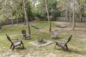 A backyard with a fire pit, four Adirondack chairs, and a hammock.