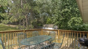 A deck with a glass-top table and chairs, surrounded by a wooden railing, overlooking a backyard with trees and a barbecue grill.