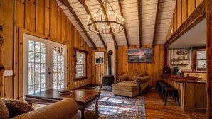 A cozy cabin interior with wooden walls, a large chandelier, a fireplace, a sofa, a coffee table, and a kitchen area. - Hike the Holler - Stay & Explore (Cosby)