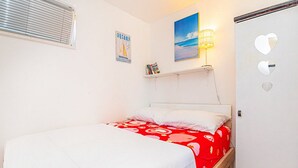 Ein Schlafzimmer mit Bett, Nachttisch, einer Wandregal mit Büchern, einem Fenster mit Jalousien und einer Tür mit Herzformen.