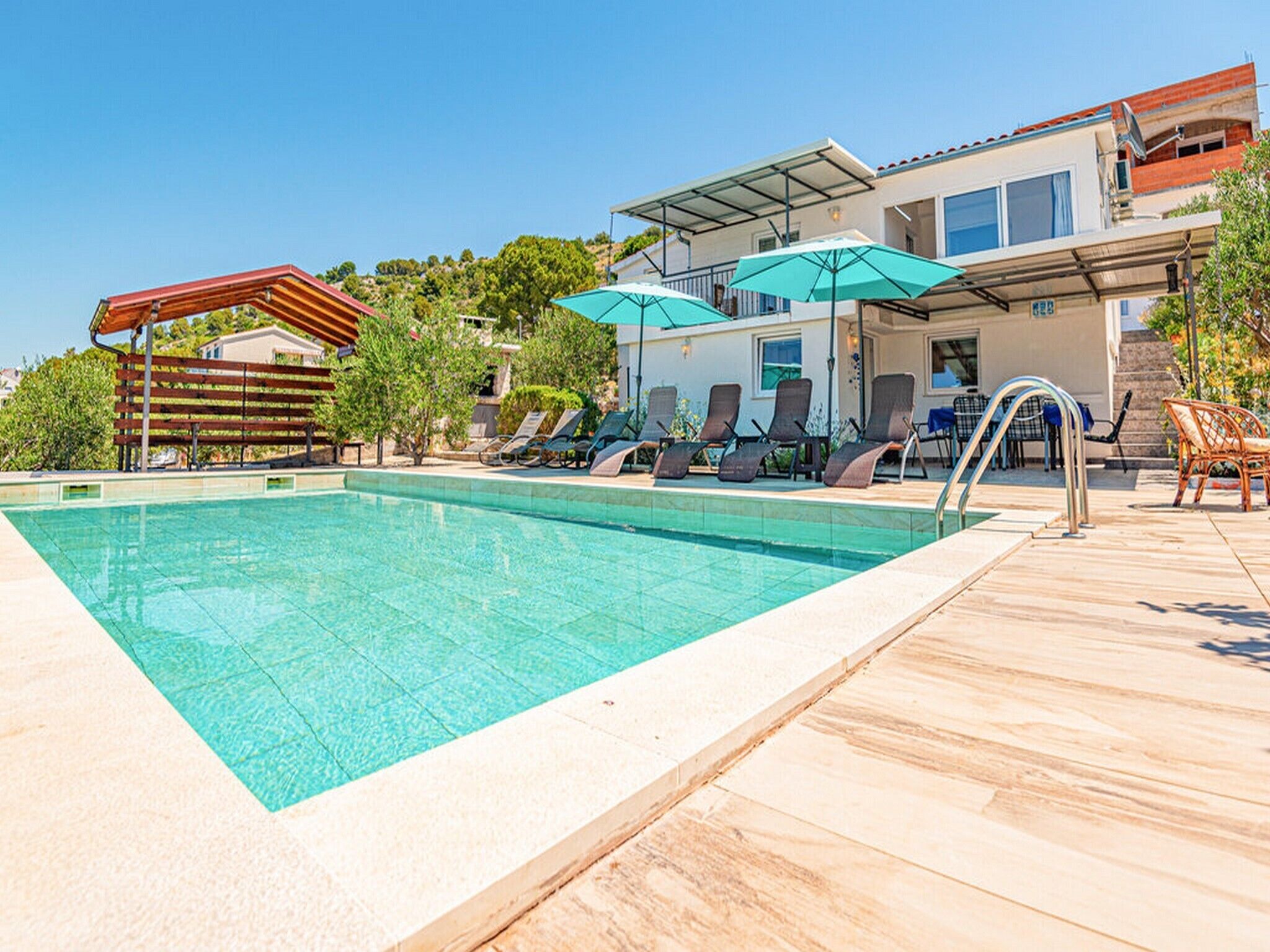 Ein Schwimmbecken mit Liegestühlen und Sonnenschirmen, umgeben von einer Holzterrasse und einem Gebäude mit überdachter Terrasse.