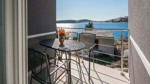 Un balcon avec une table et des chaises, un vase rempli de fleurs et une vue sur une étendue d’eau et des collines lointaines.