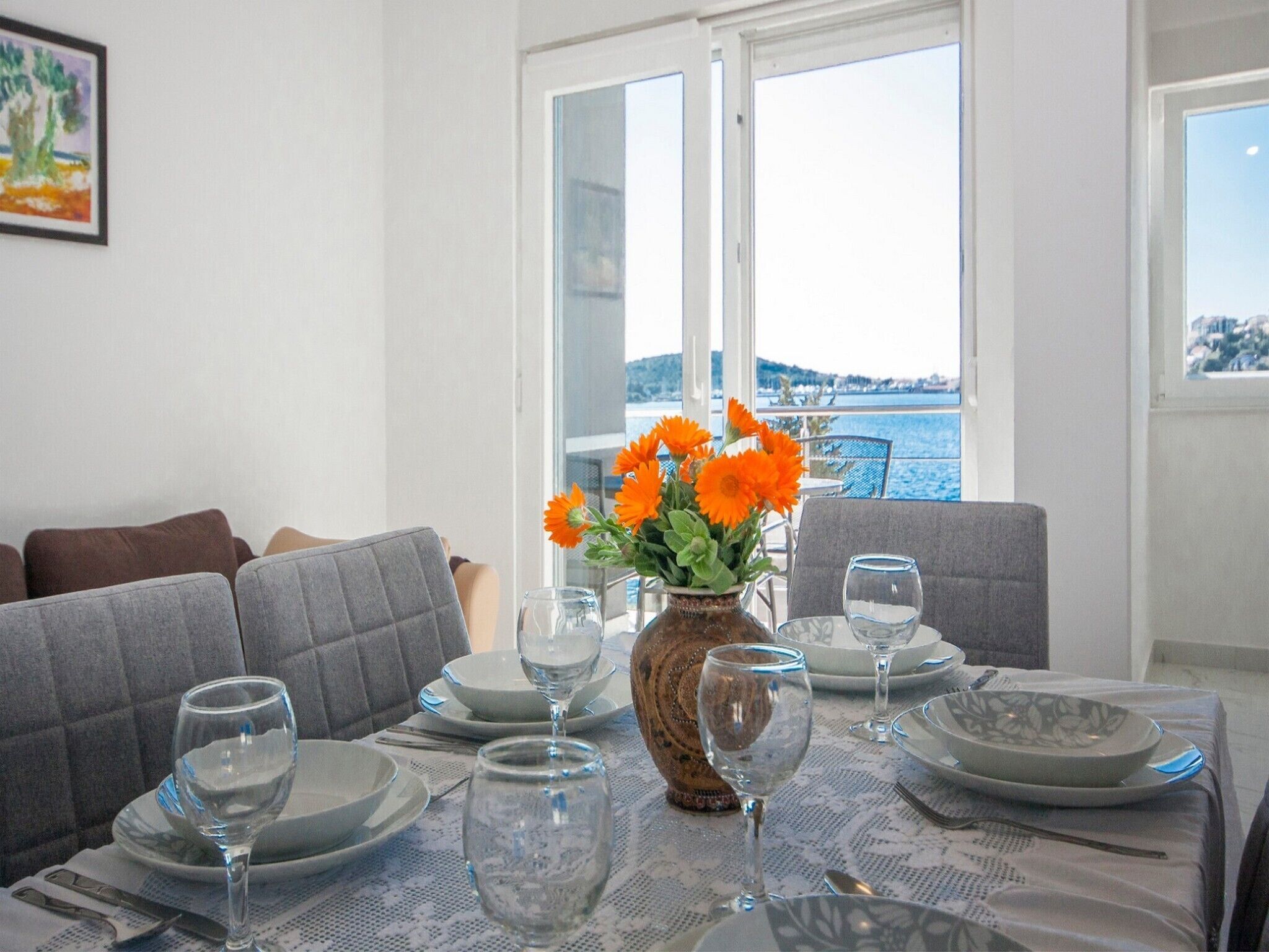 Ein Esstisch gedeckt mit Tellern, Gläsern und Besteck, eine Vase mit Blumen und Blick auf das Meer durch offene Türen.