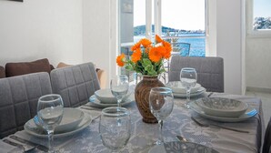 Une table à manger dressée avec des assiettes, des verres et des couverts, un vase rempli de fleurs et une vue sur la mer par des portes ouvertes.