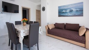 A living room with a dining area, a flat-screen TV, a clock, and a painting of the ocean. - Apartments Happy Holiday - Two Bedroom Apartment with Terrace (Rogoznica)