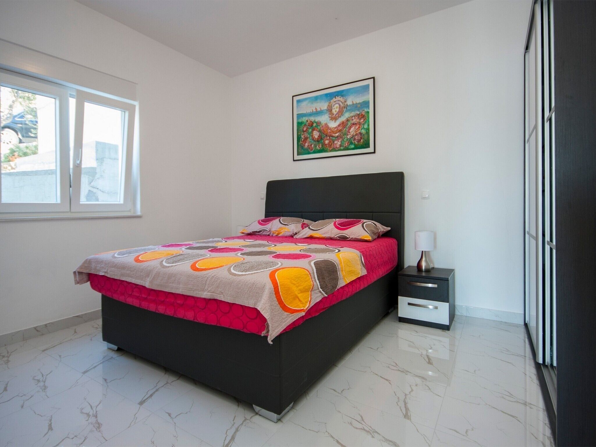 Un dormitorio con cama, mesita de noche, lámpara, ventana y un cuadro en la pared.