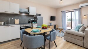 A modern kitchen with white cabinets, a dining table, and chairs. There is a sofa and a TV in the living area.