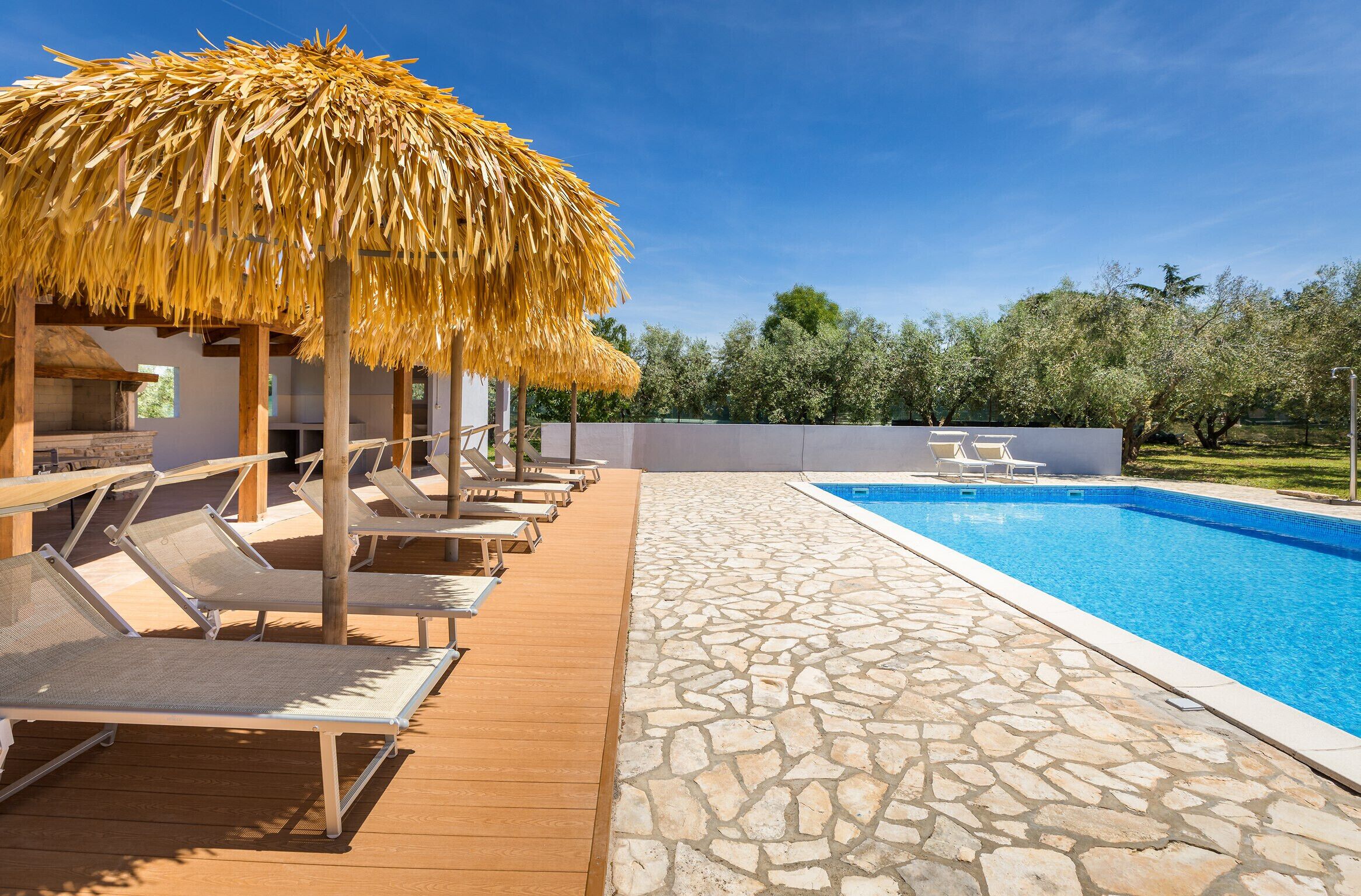 A poolside area with lounge chairs and a thatched umbrella, a stone-tiled deck, and a clear blue pool.