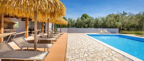 A poolside area with lounge chairs and a thatched umbrella, a stone-tiled deck, and a clear blue pool.