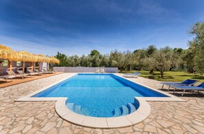 Uma piscina retangular com borda de pedra, rodeada por espreguiçadeiras e uma estrutura com telhado de palha.