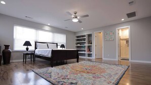 Une chambre à coucher comprenant un lit, une table de chevet, une lampe, un grand tapis, un ventilateur de plafond, une bibliothèque et une salle de bain visible à travers une porte ouverte.