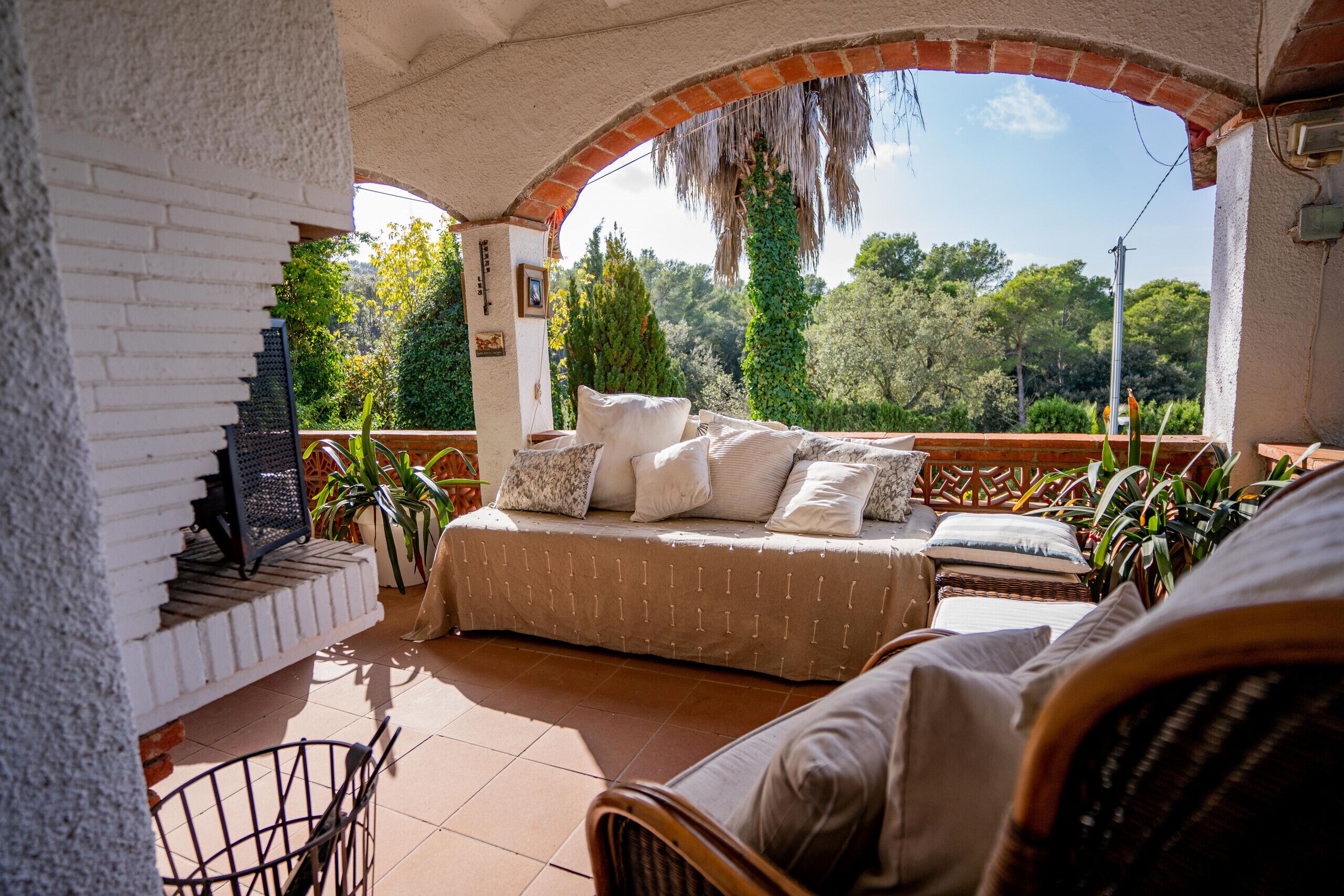 A cozy outdoor seating area with a sofa and chairs, a brick fireplace, and a view of lush greenery.