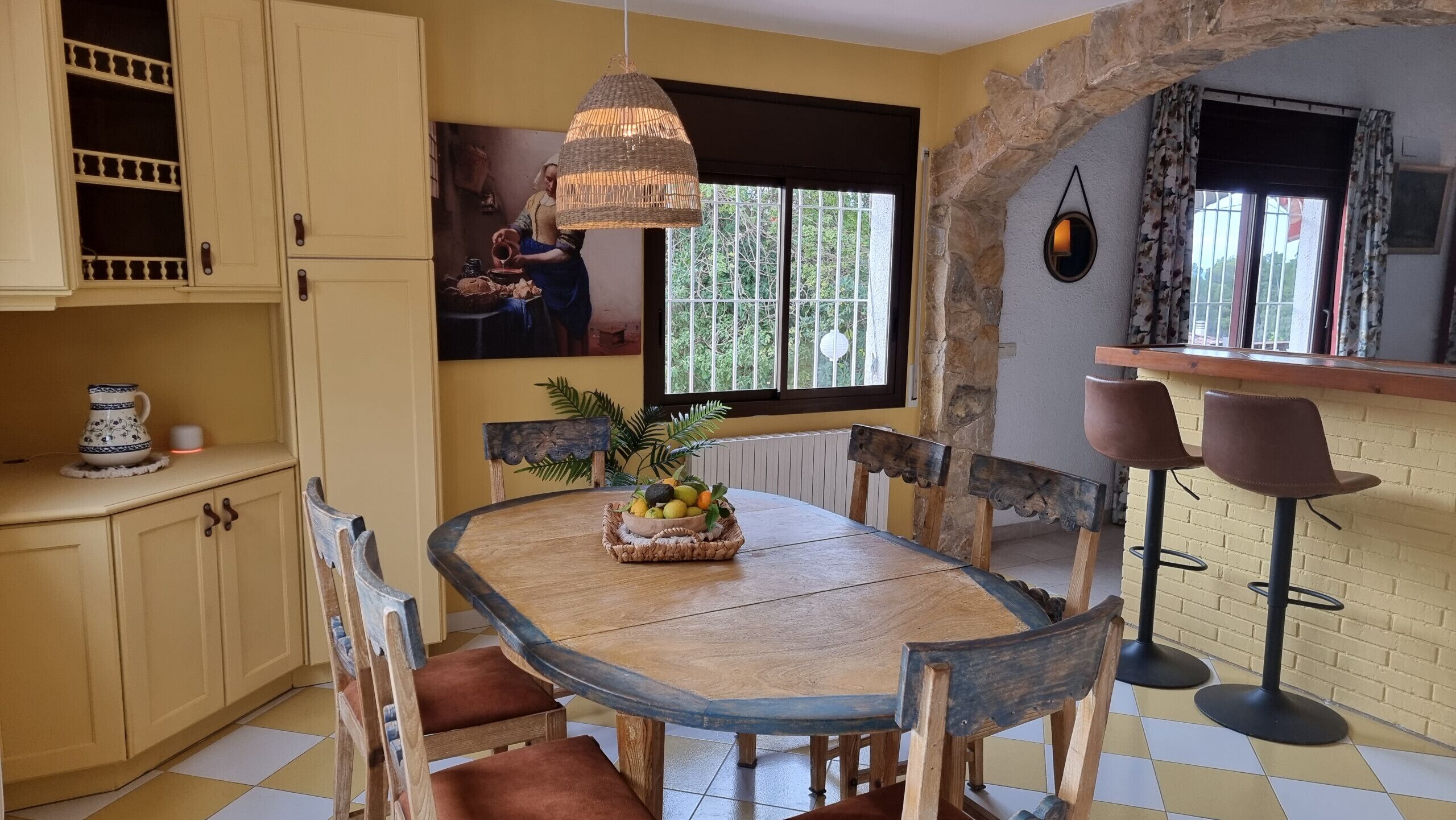 Un coin repas avec une table ronde en bois, des chaises et un panier de fruits. On y trouve une arche en pierre, une fenêtre avec vue et un luminaire suspendu.
