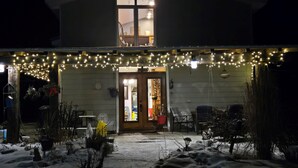 Una casa con porche cubierto decorado con luces colgantes, una puerta que da a un interior con comedor, y nieve en el suelo.
