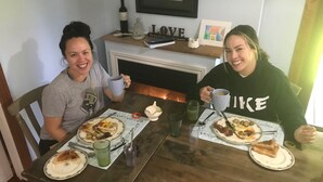 Dos personas sentadas en una mesa de madera con platos de comida, vasos y una botella de vino.