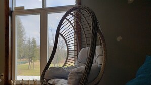 Una habitación con un ventanal grande, una silla colgante de mimbre y un cojín azul.