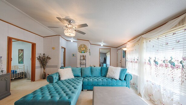 A living room with a teal sectional sofa, a matching ottoman, and a white coffee table. There is a ceiling fan, a chandelier, and a window with floral curtains.