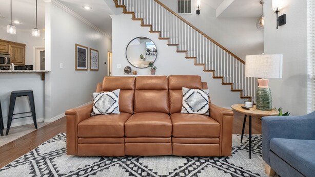 A living room with a brown leather sofa, a blue armchair, a patterned rug, a wooden coffee table, and a staircase with white railings. - The West Nest 2 -Families, 2 Kings, Game Room (Canyon)