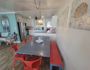 Un coin repas avec une longue table, des chaises rouges et un banc. On aperçoit une cuisine en arrière-plan, avec des meubles blancs et un réfrigérateur.