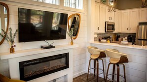 Una cocina moderna con armarios blancos, un televisor de pantalla plana grande montado sobre la chimenea y una zona de bar con taburetes.