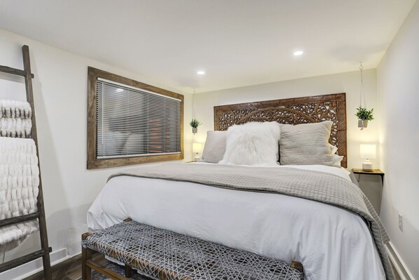 A neatly arranged bedroom with a large bed, a bench, a wooden headboard, a window with blinds, and a hanging plant.