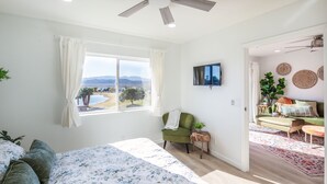 A spacious bedroom with a large bed, a view of the outdoors, a green armchair, and a TV mounted on the wall. - Stunning Lake Views!! Short walk to London Bridge, Lake, Restaurants, and Shops! (Lake Havasu City)