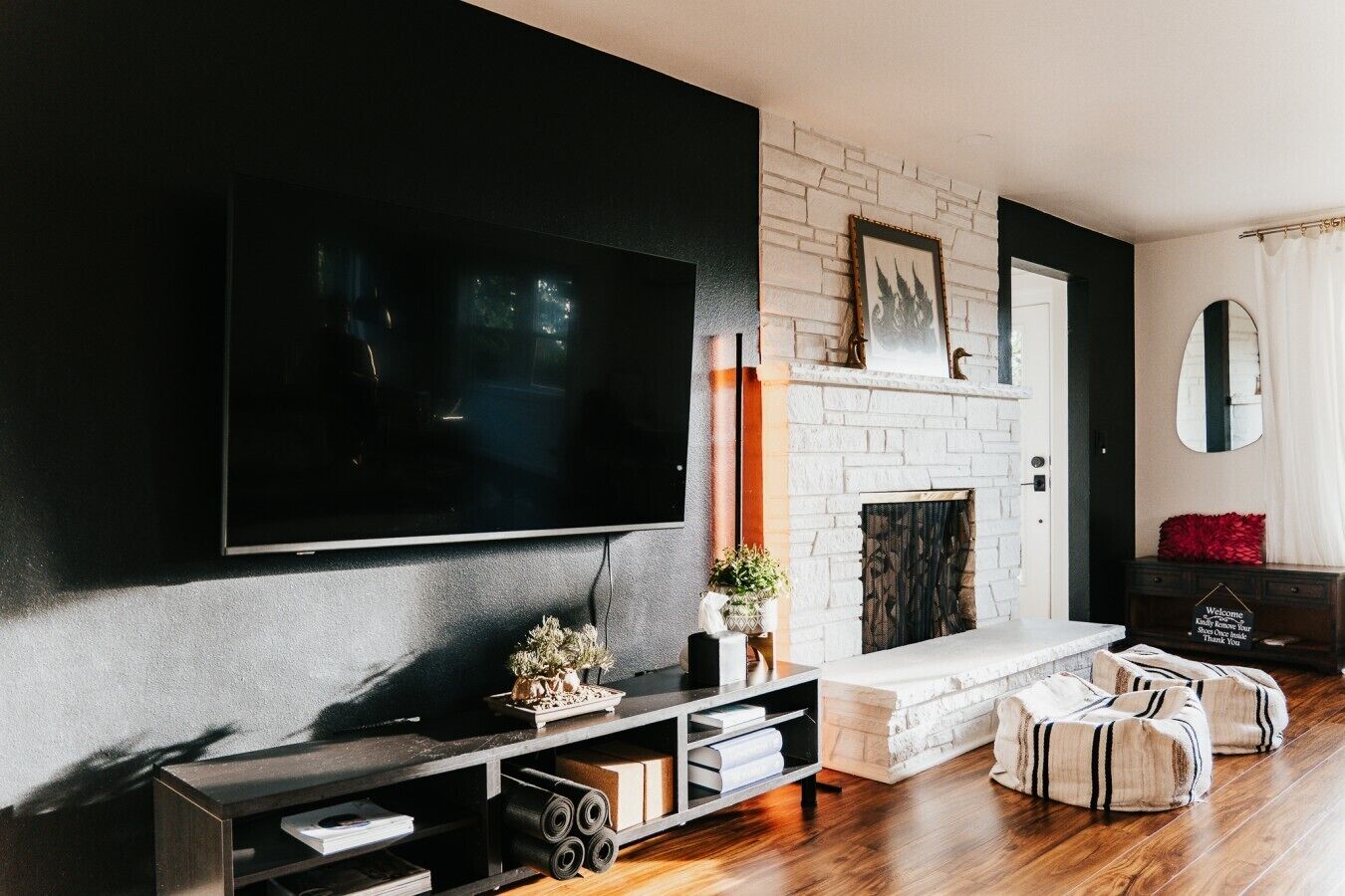 A modern living room with a large flat-screen TV mounted on the wall, a fireplace with a white brick surround, and a cozy seating area with cushions.