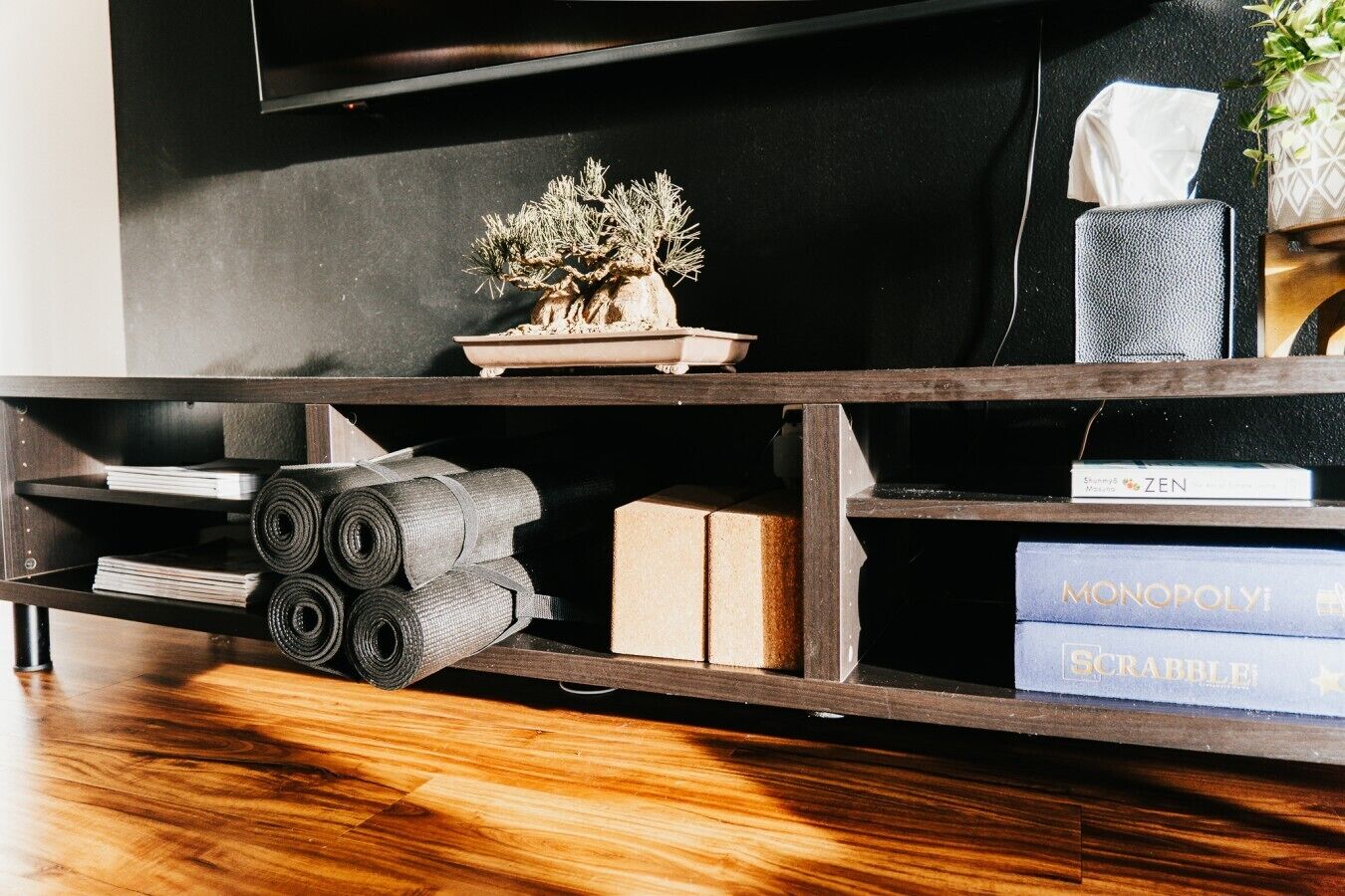 A wooden TV stand with a potted plant, rolled-up mats, and board games.