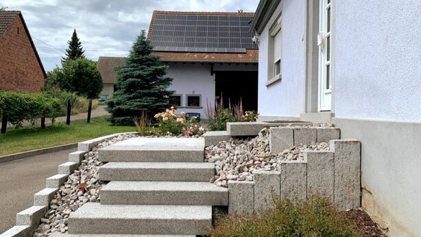 Un immeuble résidentiel doté d’un toit recouvert de panneaux solaires, d’un escalier en pierre menant à l’entrée et d’un jardin agrémenté de diverses plantes et fleurs.