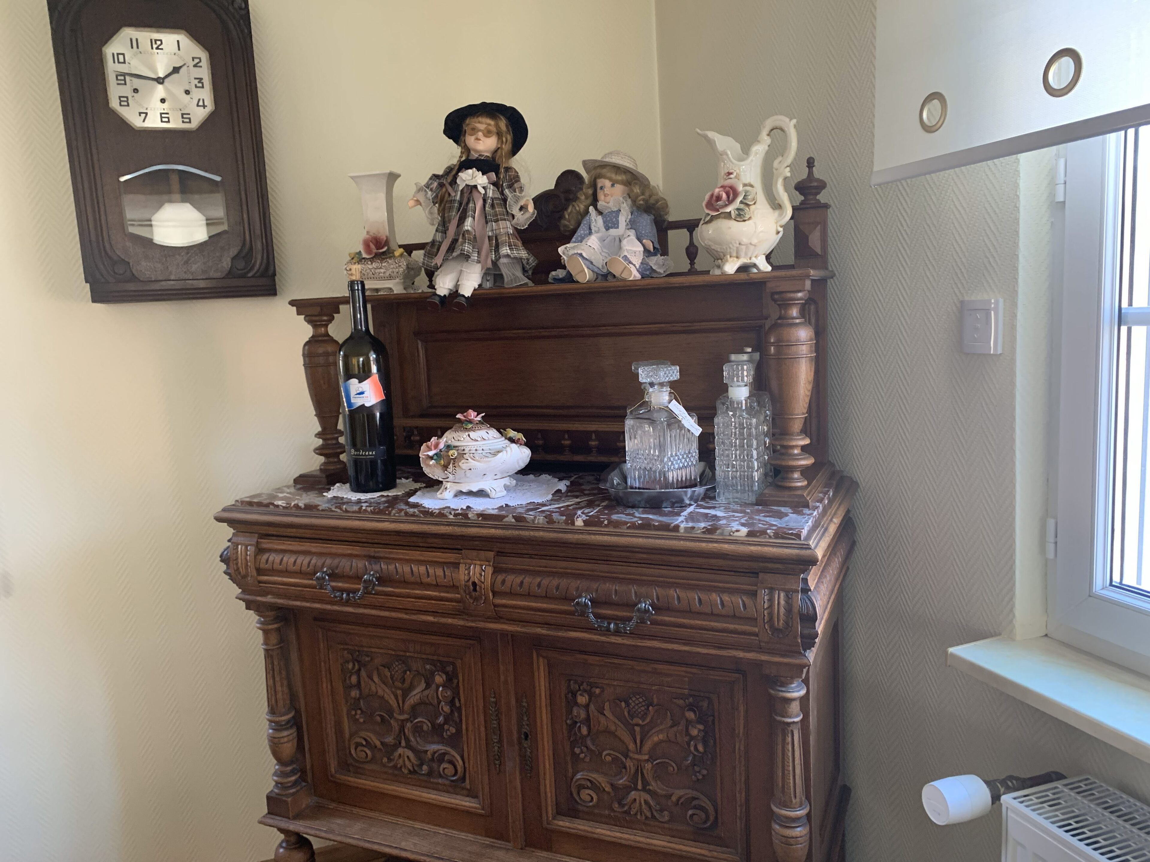 A wooden dresser with a clock above it, a bottle of wine, and decorative items on top.
