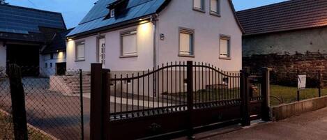 Una casa de dos pisos con techo oscuro, entrada protegida por una reja y un pequeño jardín delantero.