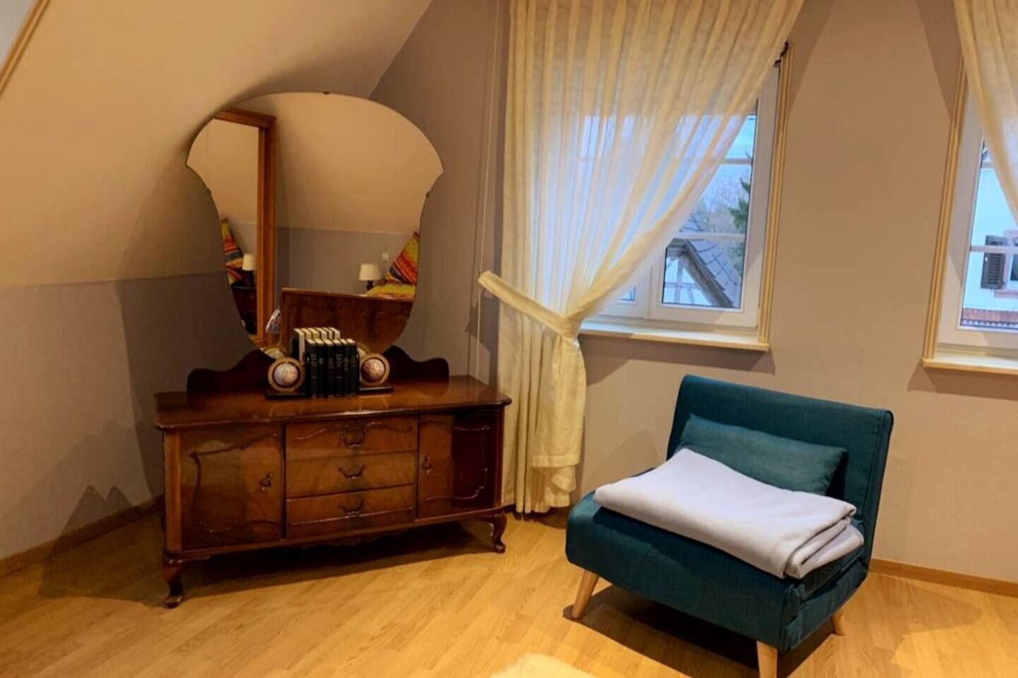 Une chambre avec une commode en bois, un fauteuil turquoise et un grand miroir.