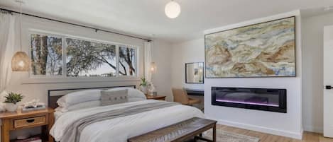 Un dormitorio moderno con una cama grande, mesitas de noche, un banco, una chimenea y obras de arte en las paredes.