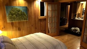 Une pièce chaleureuse aux murs en bois, un lit avec une literie à rayures, un tableau représentant des arbres et un chien qui se repose dans un coin.