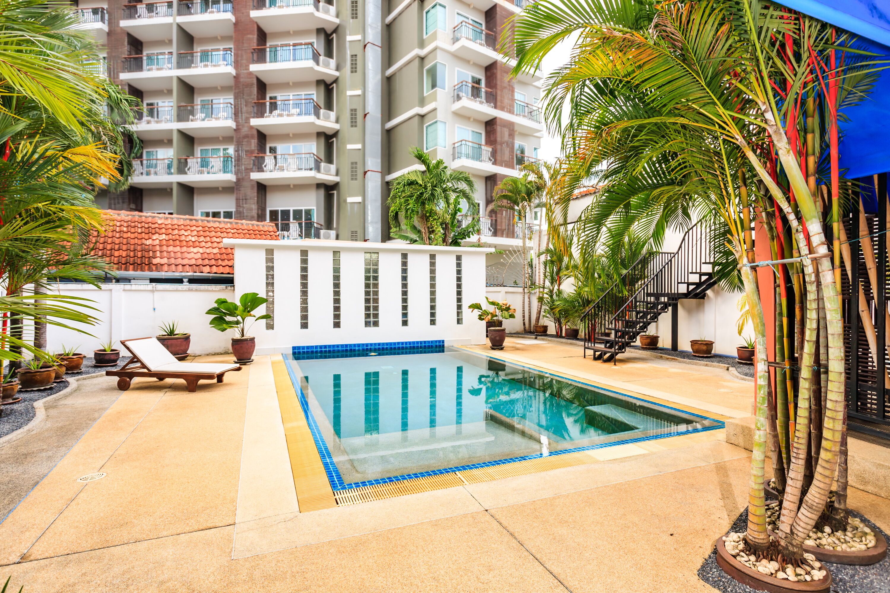 A swimming pool with lounge chairs, surrounded by potted plants and a multi-story building in the background.